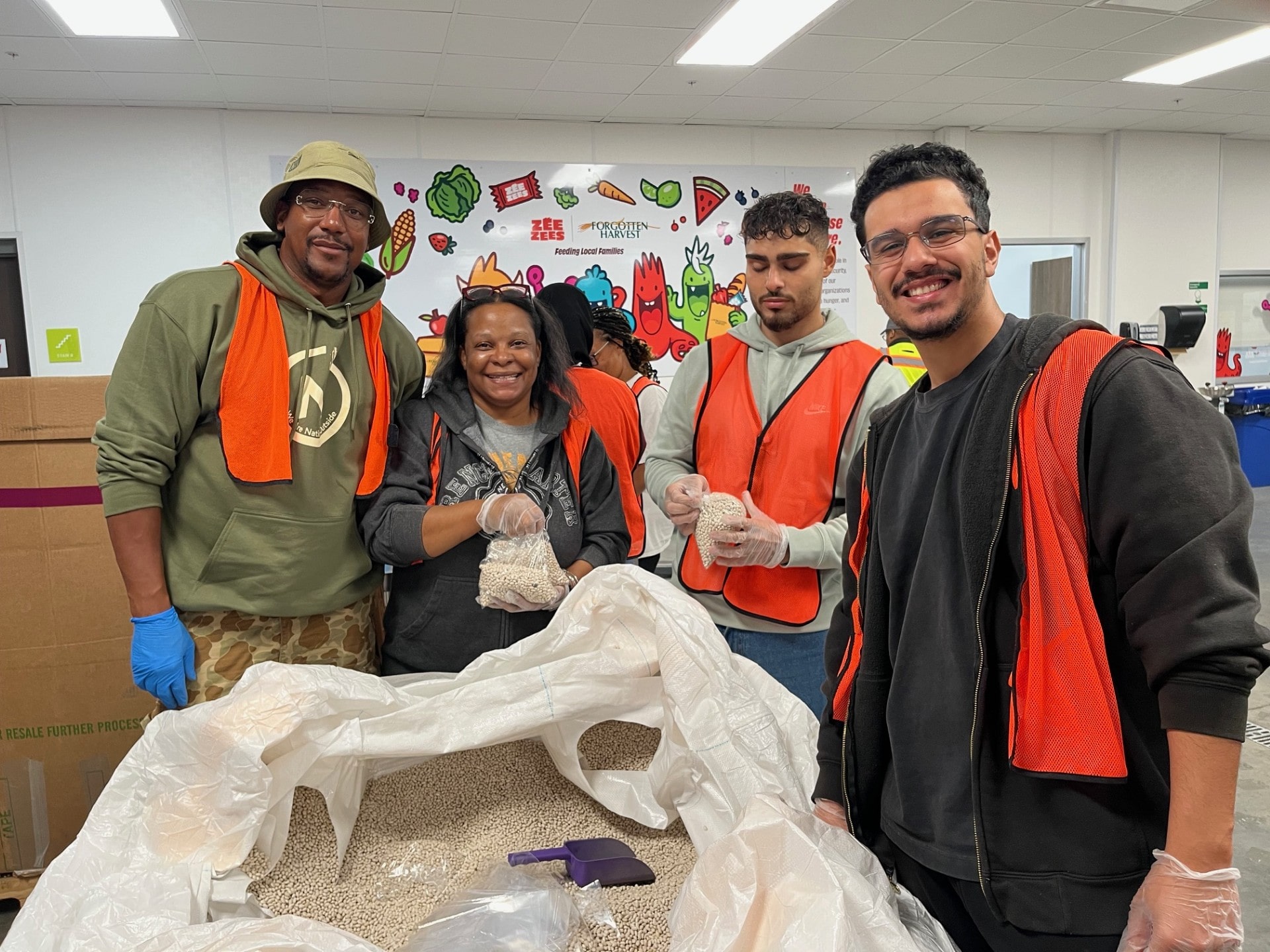 Four volunteers wearing safety vests and gloves work together at a food packaging event. They are standing behind a large bag filled with beans, in a room decorated with colorful wall art featuring fruits and vegetables.