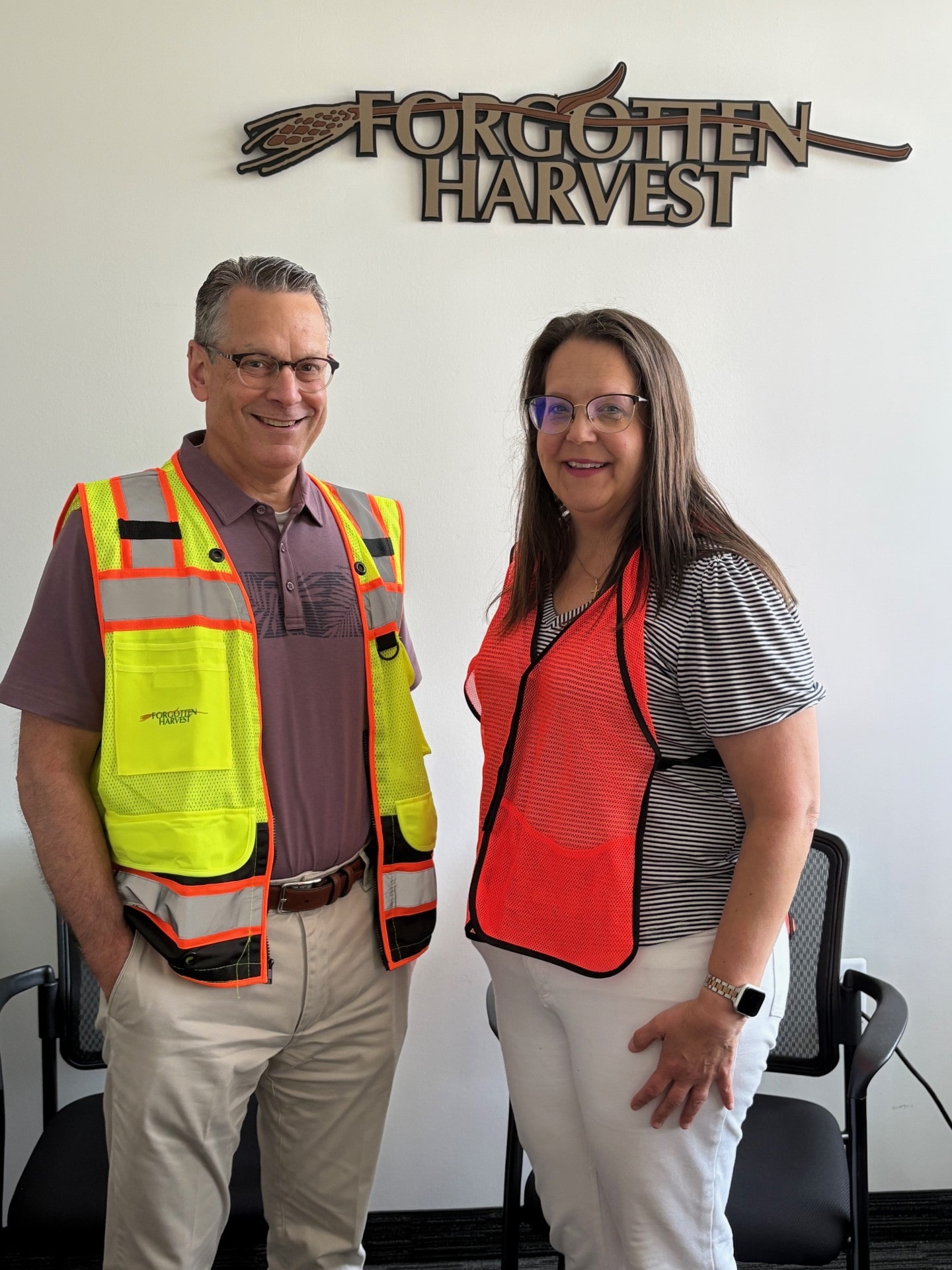Two individuals wearing safety vests stand smiling in front of a 'Forgotten Harvest' sign.