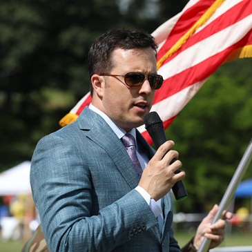 portrait of Brian Marl in a grey blazer, purple tie and sunglasses, holding an American flag