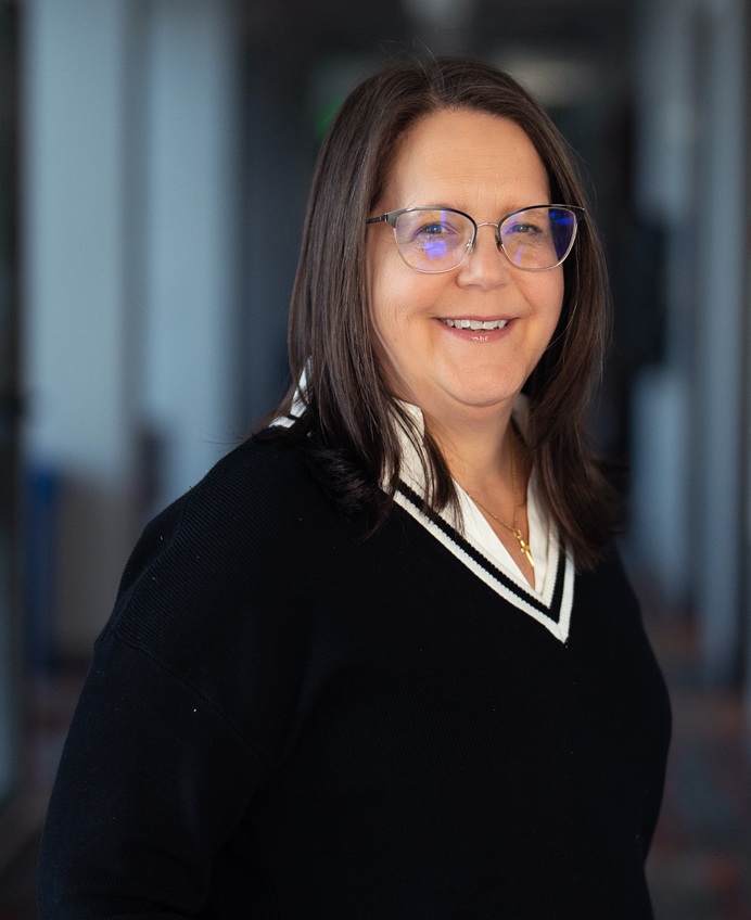 A person with shoulder‑length brown hair stands indoors wearing a black V‑neck sweater with a white trim