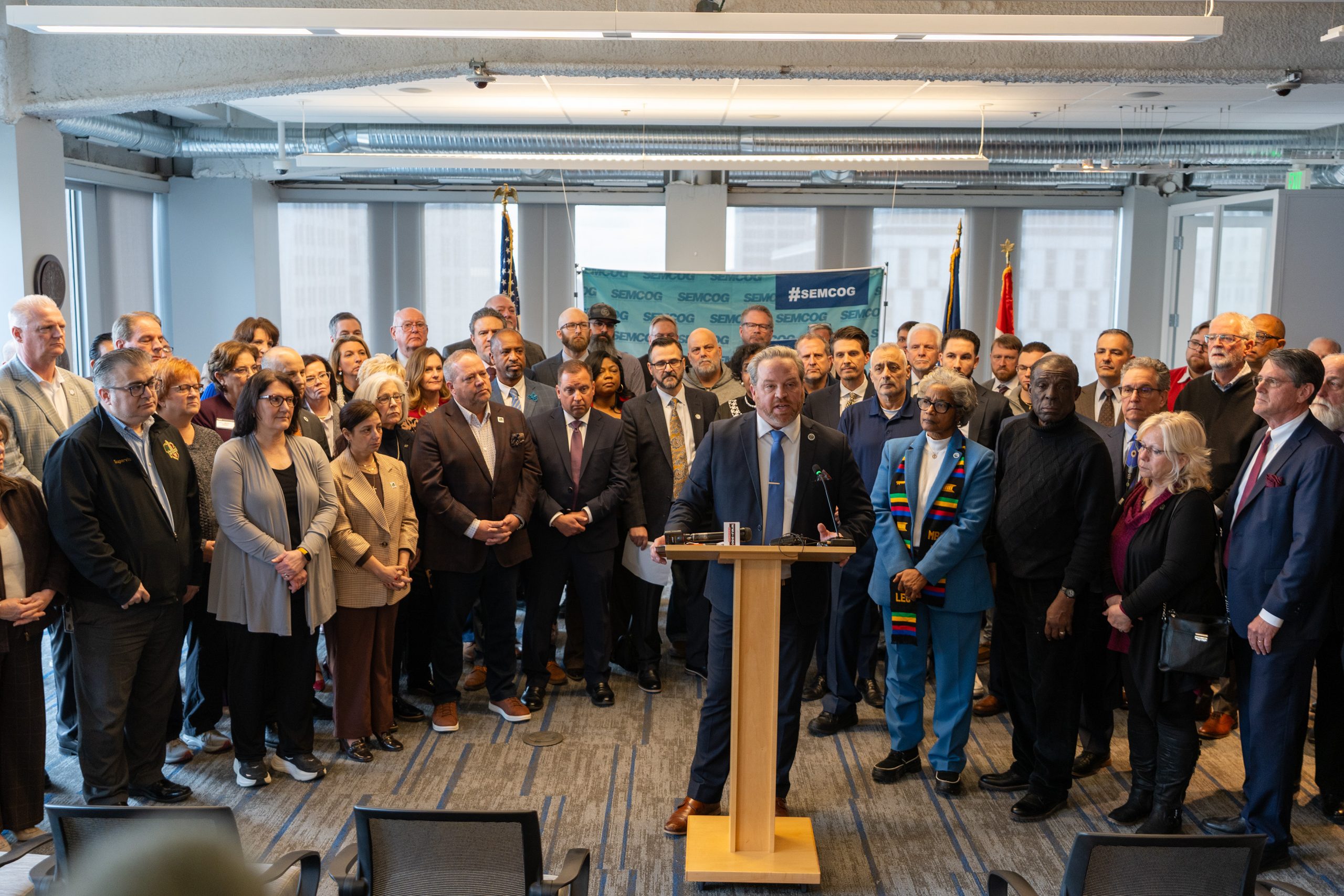 large group of people standing behind podium where man in suit and tie is speaking