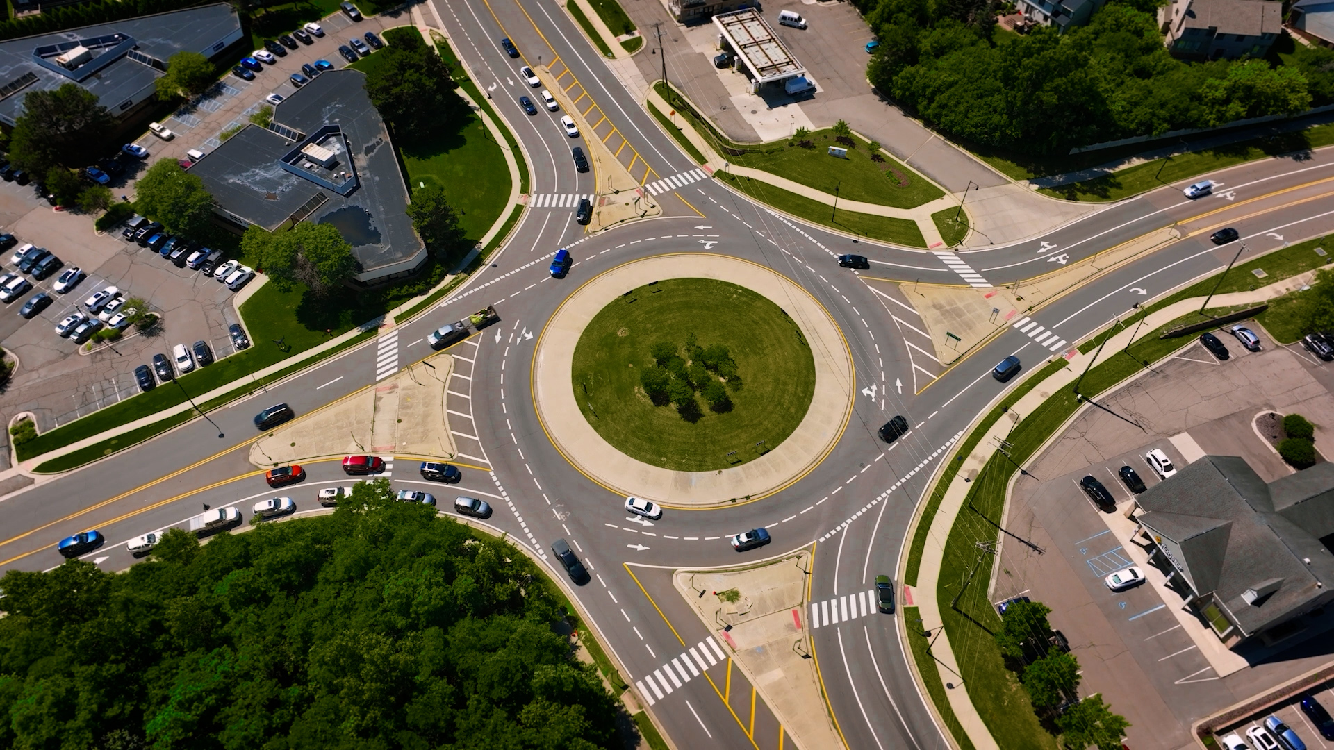 Aerial view of a large multi‑lane roundabout surrounded by roads, crosswalks, cars, parking lots, and trees, with landscaped greenery in the center of the roundabout.
