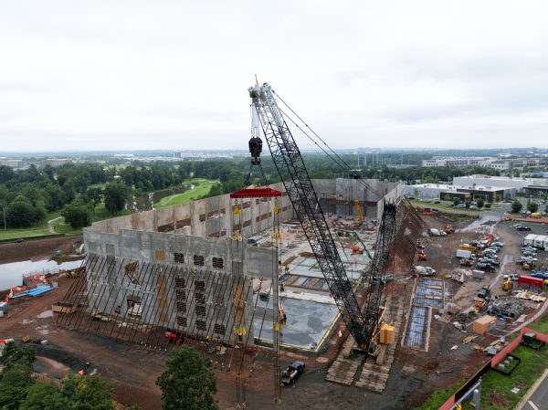 Aerial view of a data center construction site, with a large crane installing gray structural walls around the edges of the foundation and various trucks, construction trailers, and equipment staged around the site.