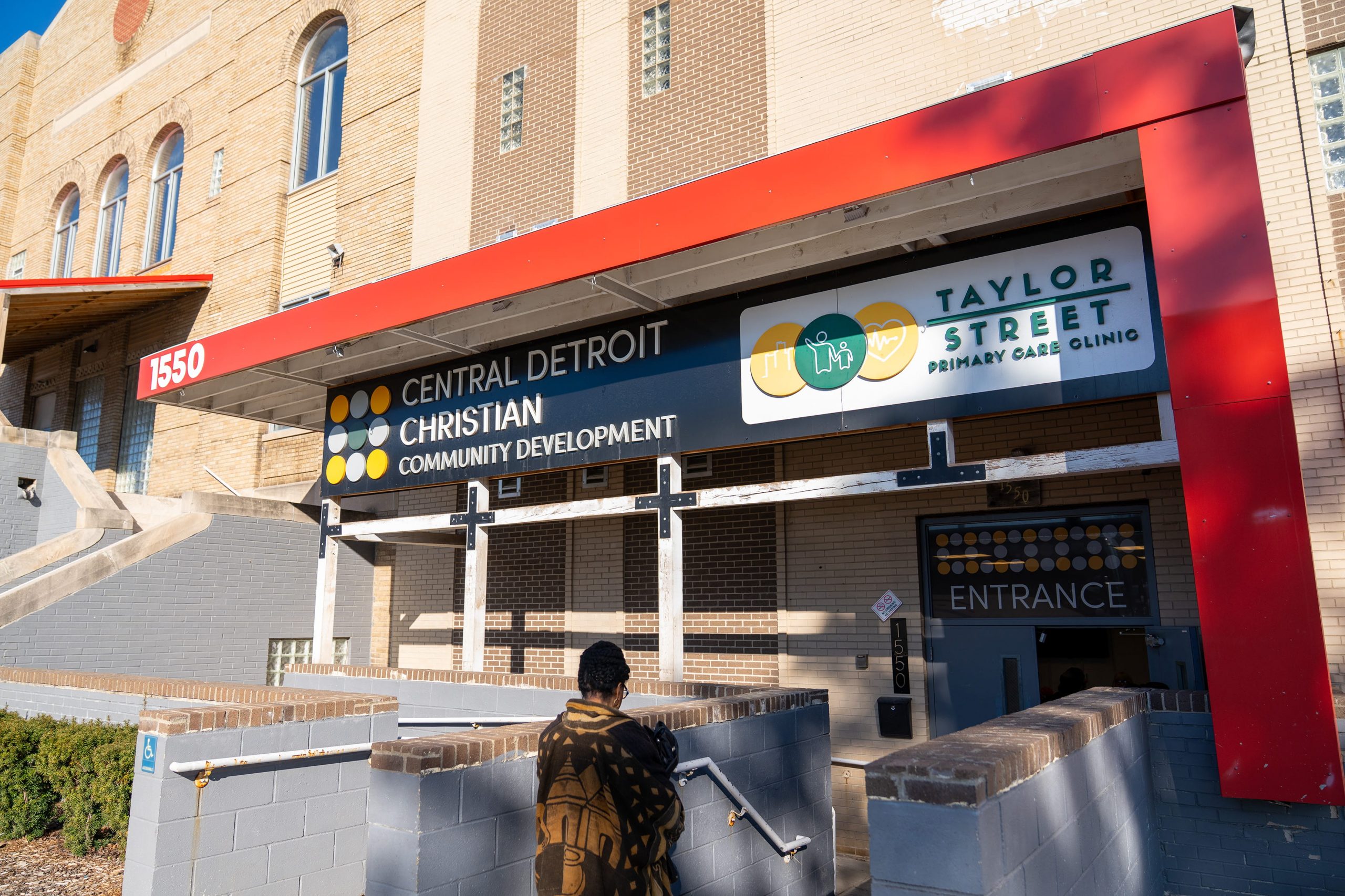 entrance to a building housing Central Detroit Christian Community Development and Taylor Street Primary Care Clinic, featuring a red awning with address number 1550. Signage includes logos and text in white, black, yellow, and teal colors, with a person walking down stairs toward the entrance.