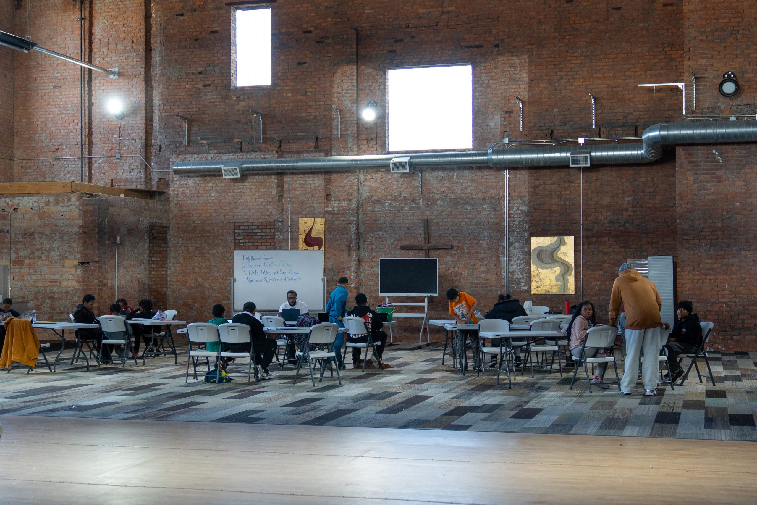 large, open indoor space with exposed brick walls and high windows, set up for a group meeting or workshop. Multiple people are seated at tables arranged in rows, with a whiteboard and large screen at the front, indicating an educational or collaborative event.