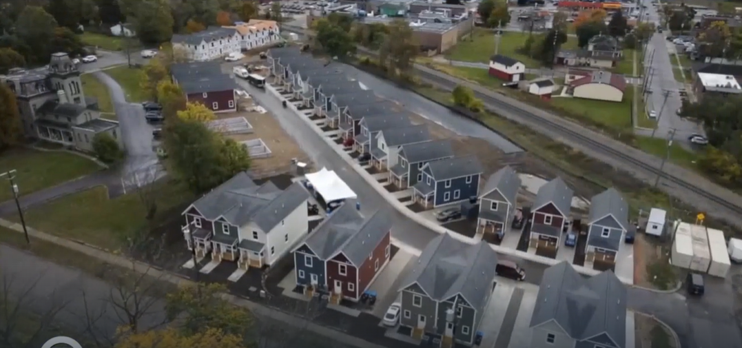 Aerial view of a newly built residential neighborhood with rows of townhomes and houses, streets, green spaces, and nearby buildings.