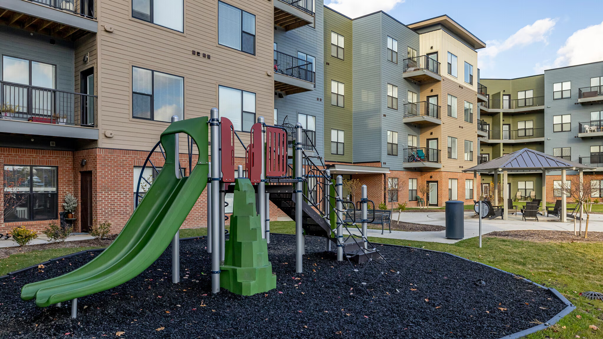 Playground with slides and climbing equipment in an apartment courtyard, surrounded by walkways, seating areas, and multi‑story residential buildings.