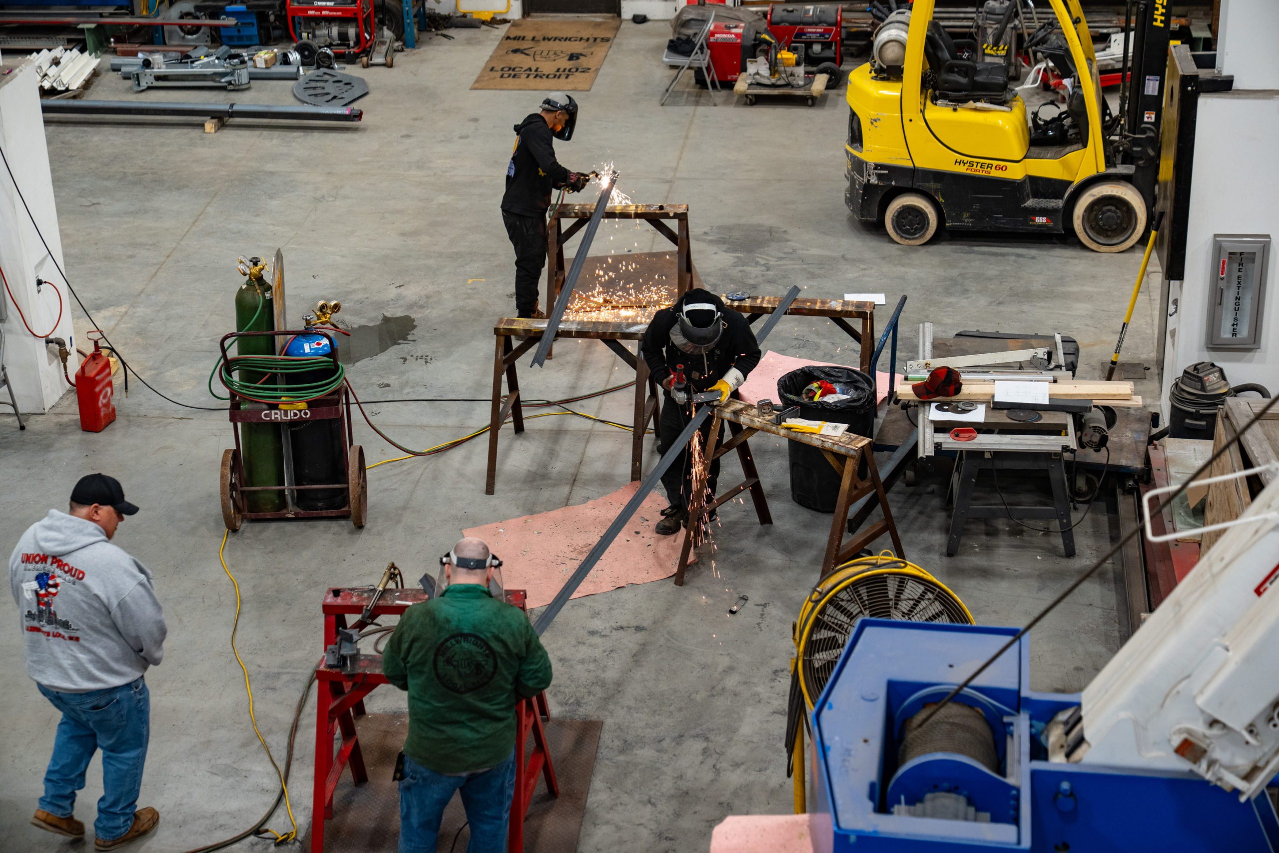 workshop with four workers engaged in metal fabrication activities, including cutting and welding steel beams. The scene features various equipment such as a yellow forklift, welding machines, gas tanks, and workbenches, highlighting an active industrial environment focused on construction or manufacturing tasks.