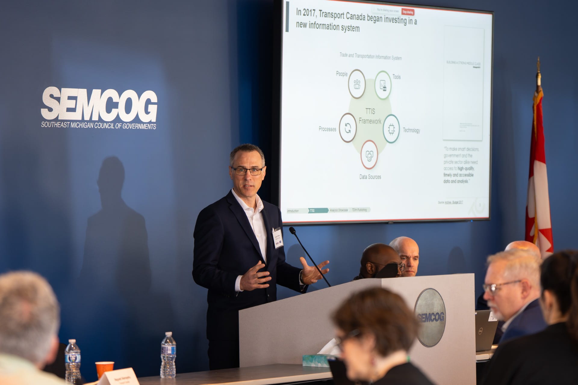 A person is speaking at a podium in a conference room with a 'SEMCOG' logo on the wall. A presentation slide is displayed, discussing Transport Canada's investment in a new information system. There is a diverse group of people seated at tables, and a Canadian flag is visible in the background.