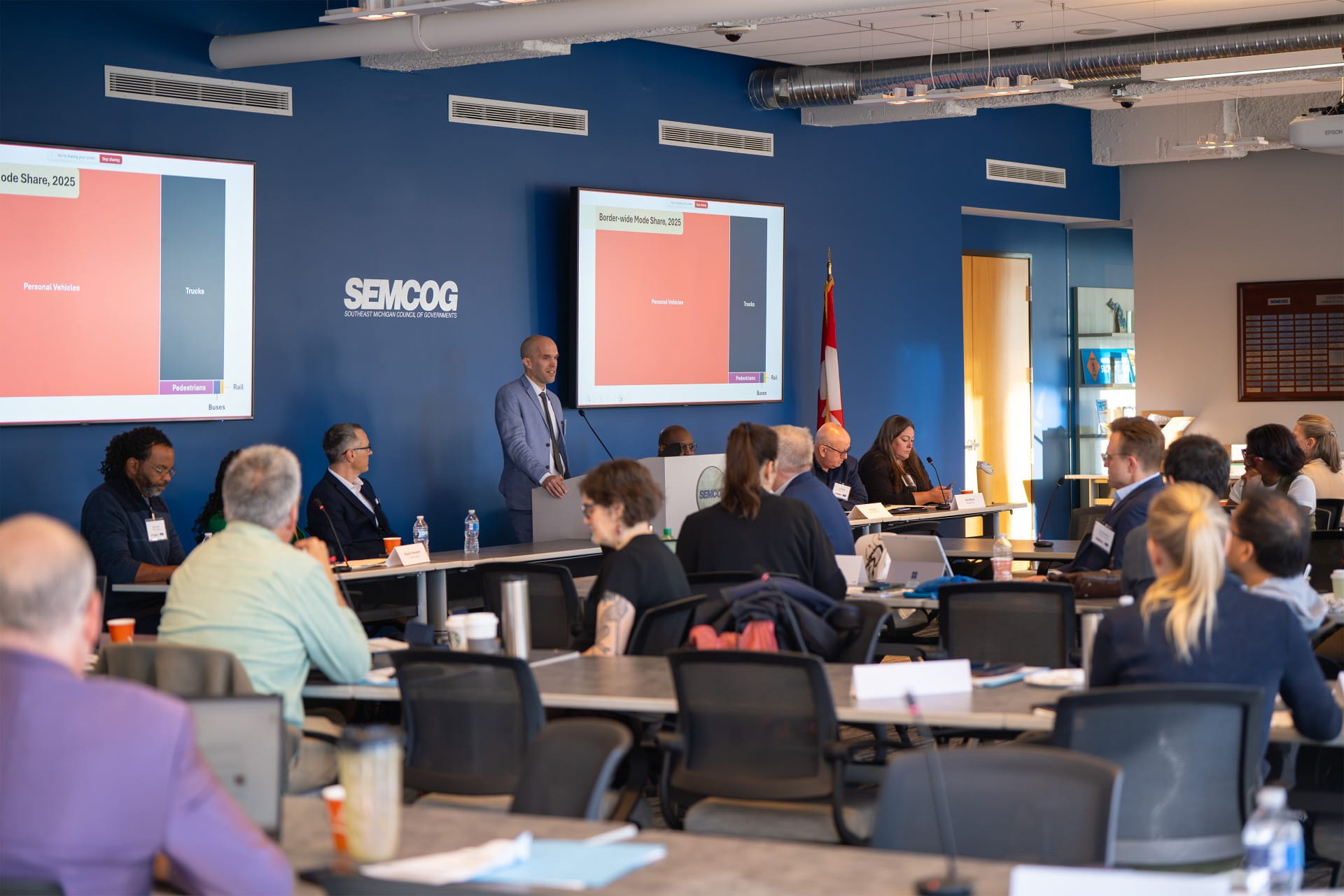 A group of people are attending a meeting in a conference room. There is a speaker standing at a podium with presentation slides displayed on two screens, one slide reading 'SEMCOG.' Attendees are seated around tables with some taking notes. An American flag is visible near the speaker.