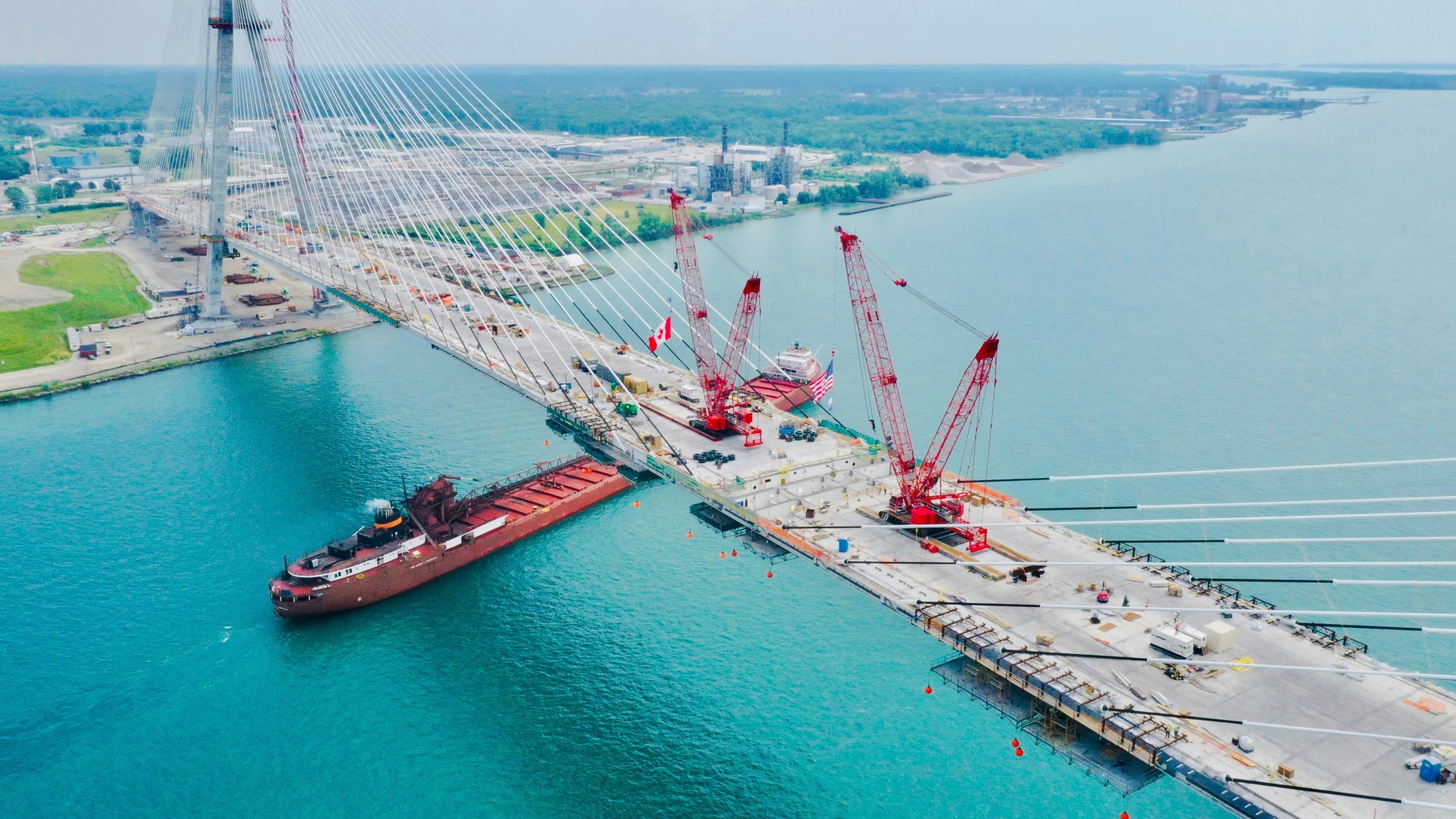Aerial view of the Gordie Howe International Bridge under construction over a wide body of water. Red cranes are positioned on the bridge deck, with a large cargo ship passing underneath. The background shows an industrial area and greenery.