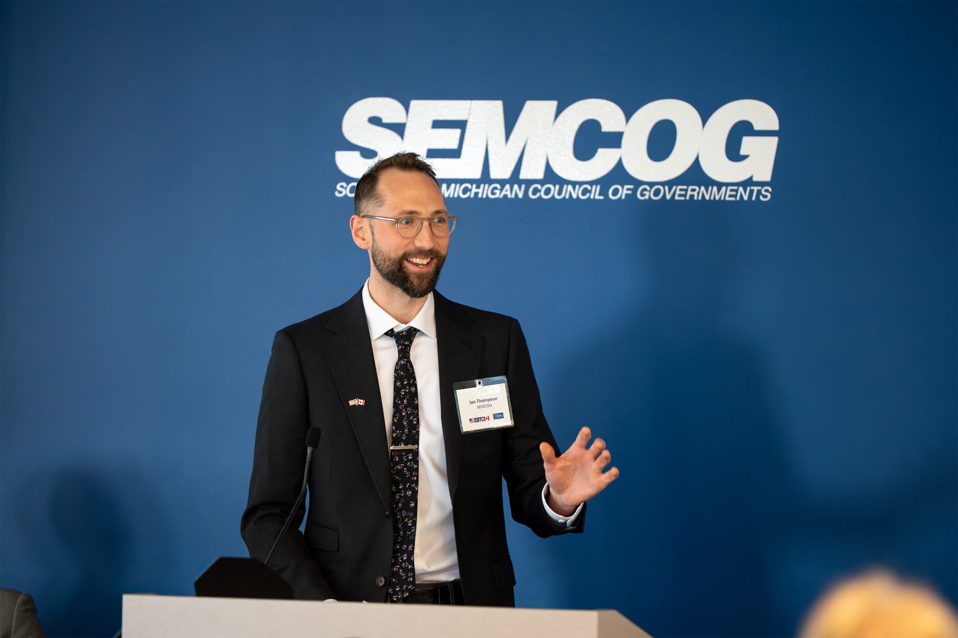A person is speaking at a podium in a conference room with a 'SEMCOG' logo on the wall.