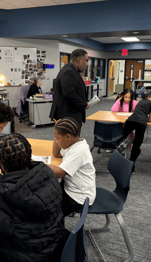 A classroom scene with several people engaged in various activities. A person in a suit stands holding papers, observing a table where young individuals are working on assignments. The room features modern furniture and educational materials.
