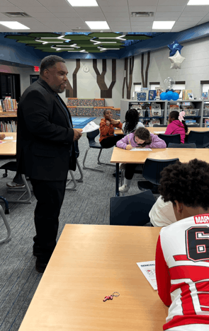 Man standing in a classroom of kids, as they are sitting at tables writing.