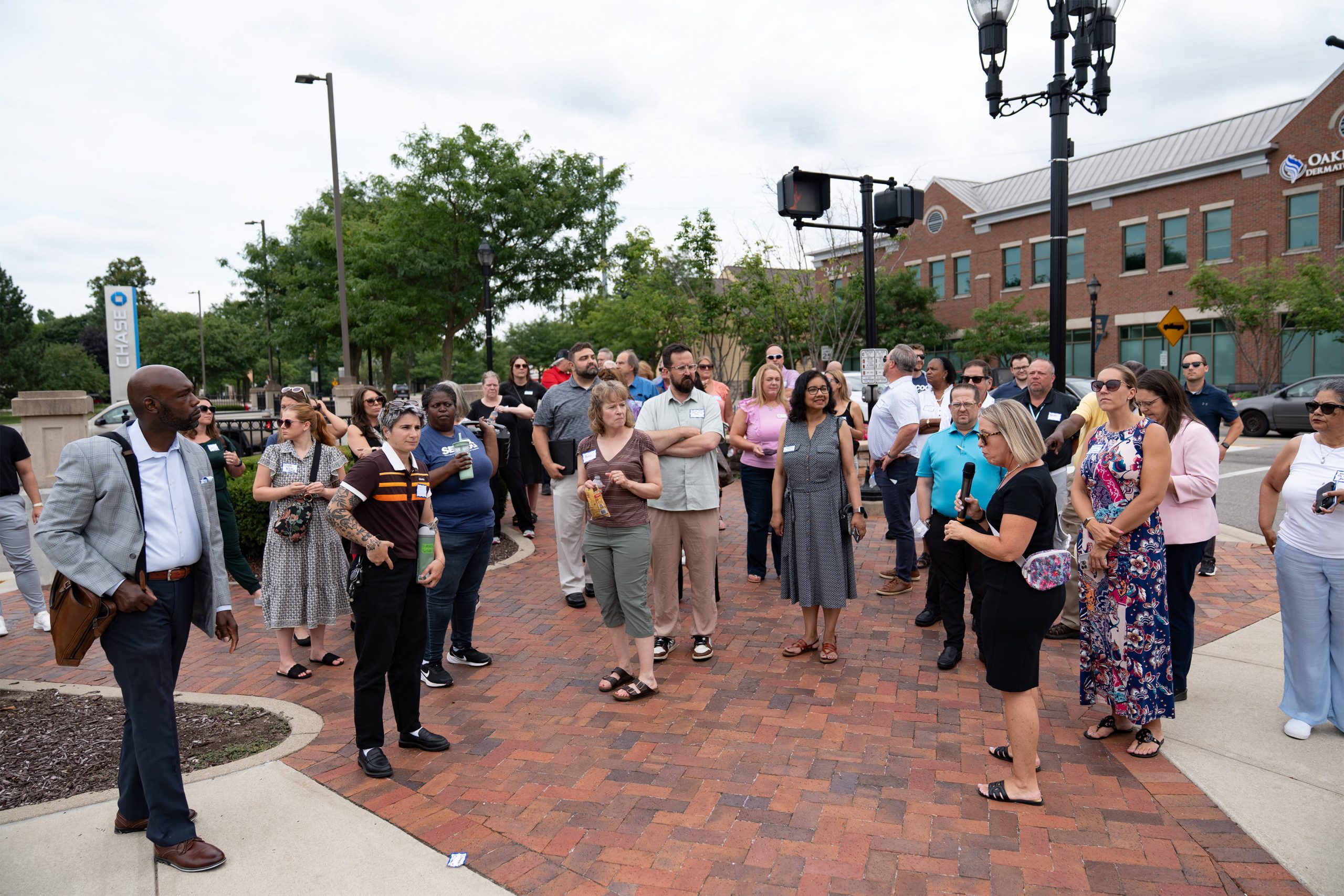 A group of people gathered on a brick-paved sidewalk, some holding drinks and engaged in conversation. In the background, there's a brick building with a sign reading 'Chick-fil-A.' Trees and lampposts are visible, suggesting an outdoor urban setting.
