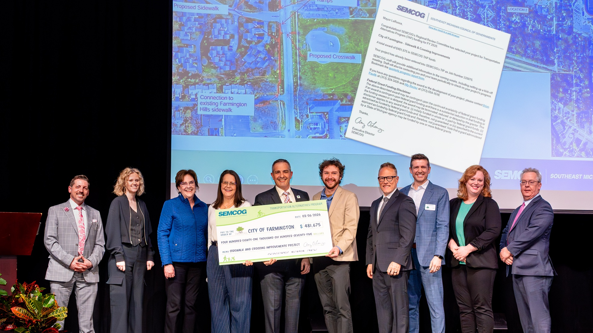 a group of 10 standing on a stage, holding a large check made out to the city of farmington