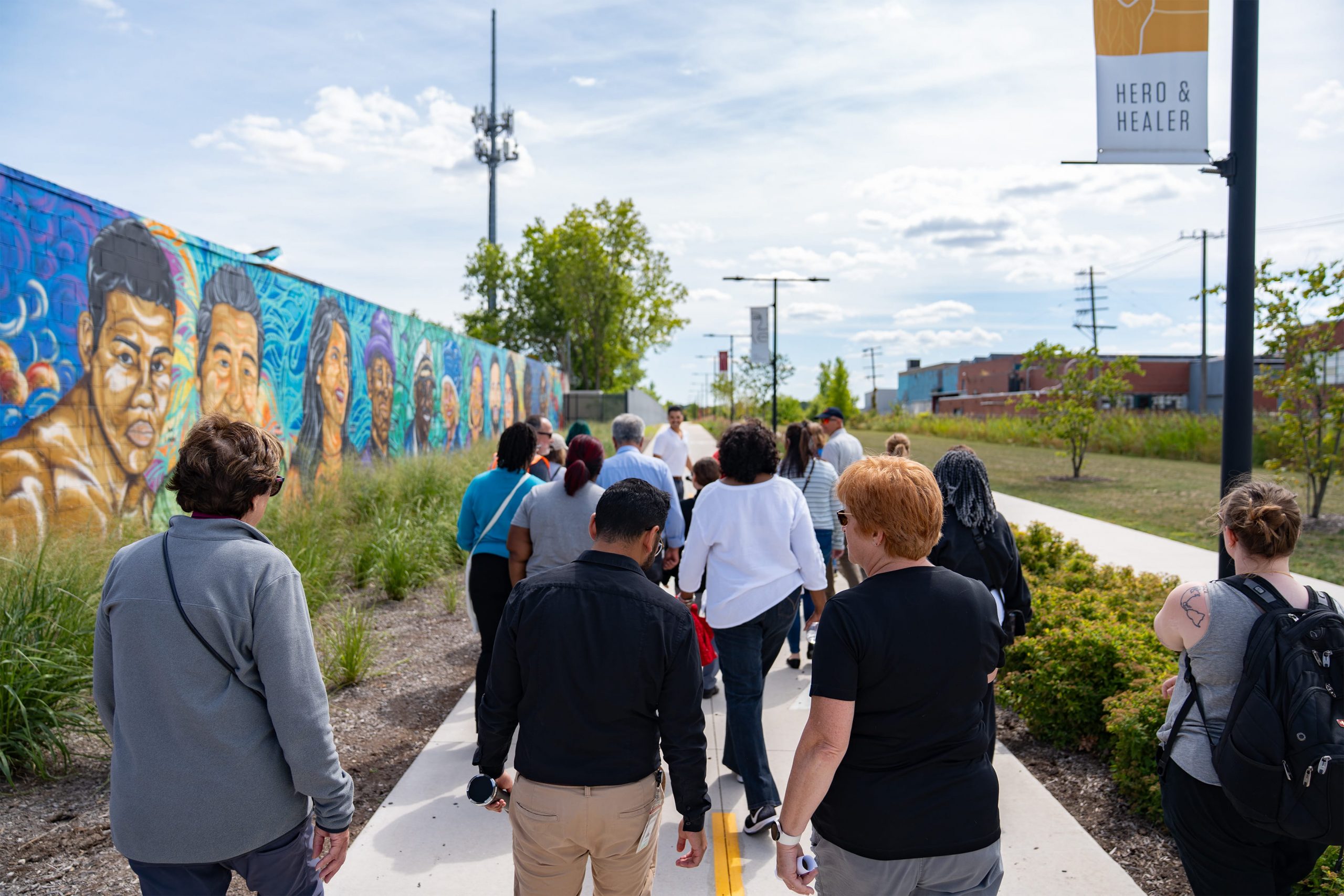 A group of people gathered on a brick-paved sidewalk, some holding drinks and engaged in conversation. In the background, there's a brick building with a sign reading 'Chick-fil-A.' Trees and lampposts are visible, suggesting an outdoor urban setting.