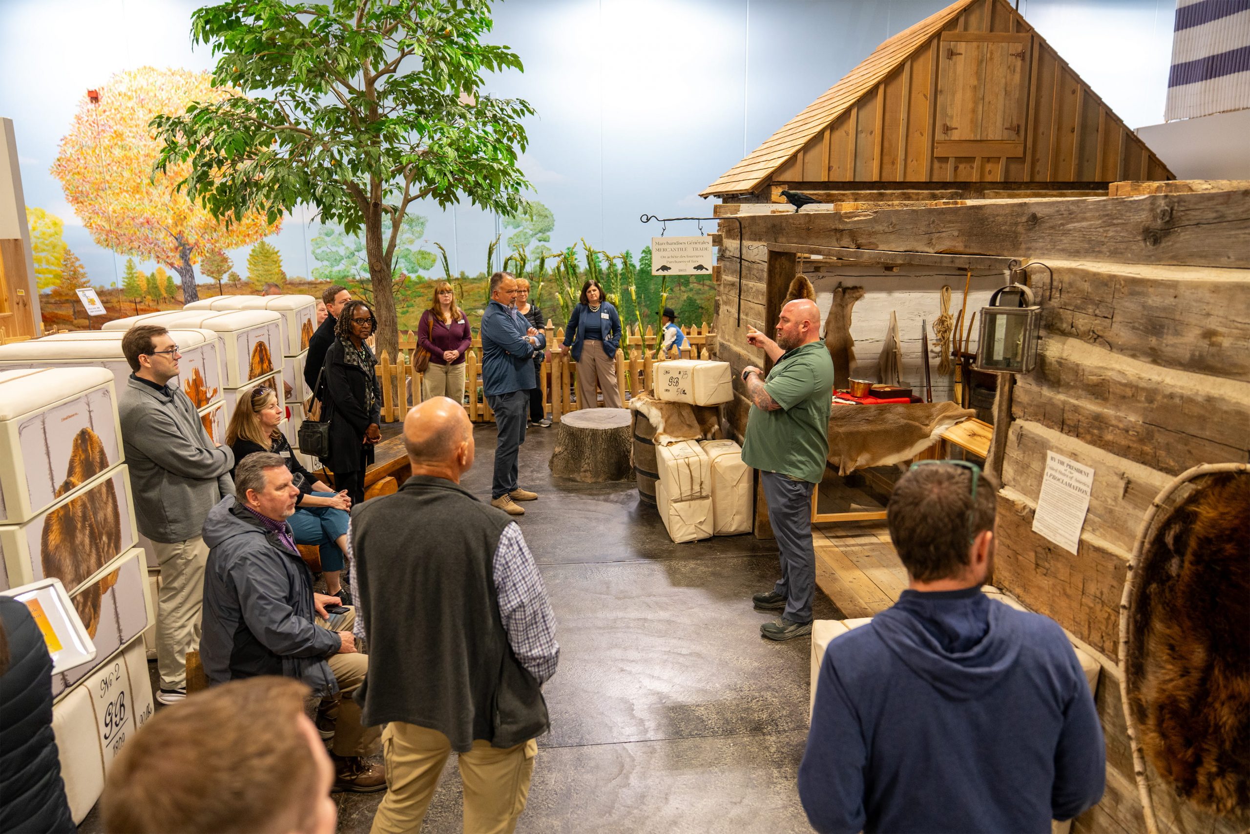A group of people gathers inside the River Raisin National Battlefield Park, listening to a speaker. The exhibit features a rustic wooden cabin and various artifacts. The atmosphere seems educational and engaging.