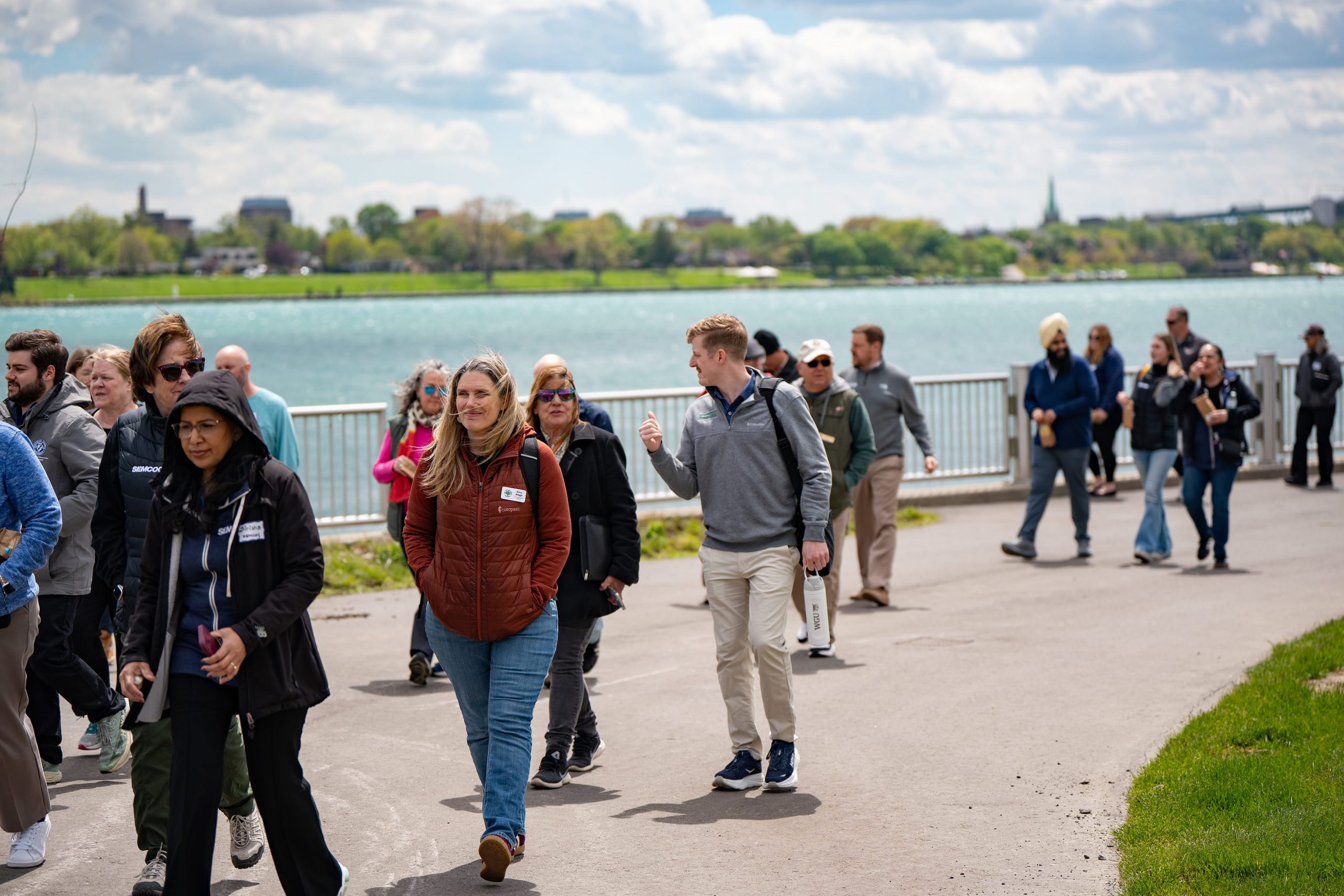 A group of people walking along a riverside path under a cloudy sky. Some individuals are wearing casual jackets and sunglasses. The background features a river with buildings visible on the opposite shore.