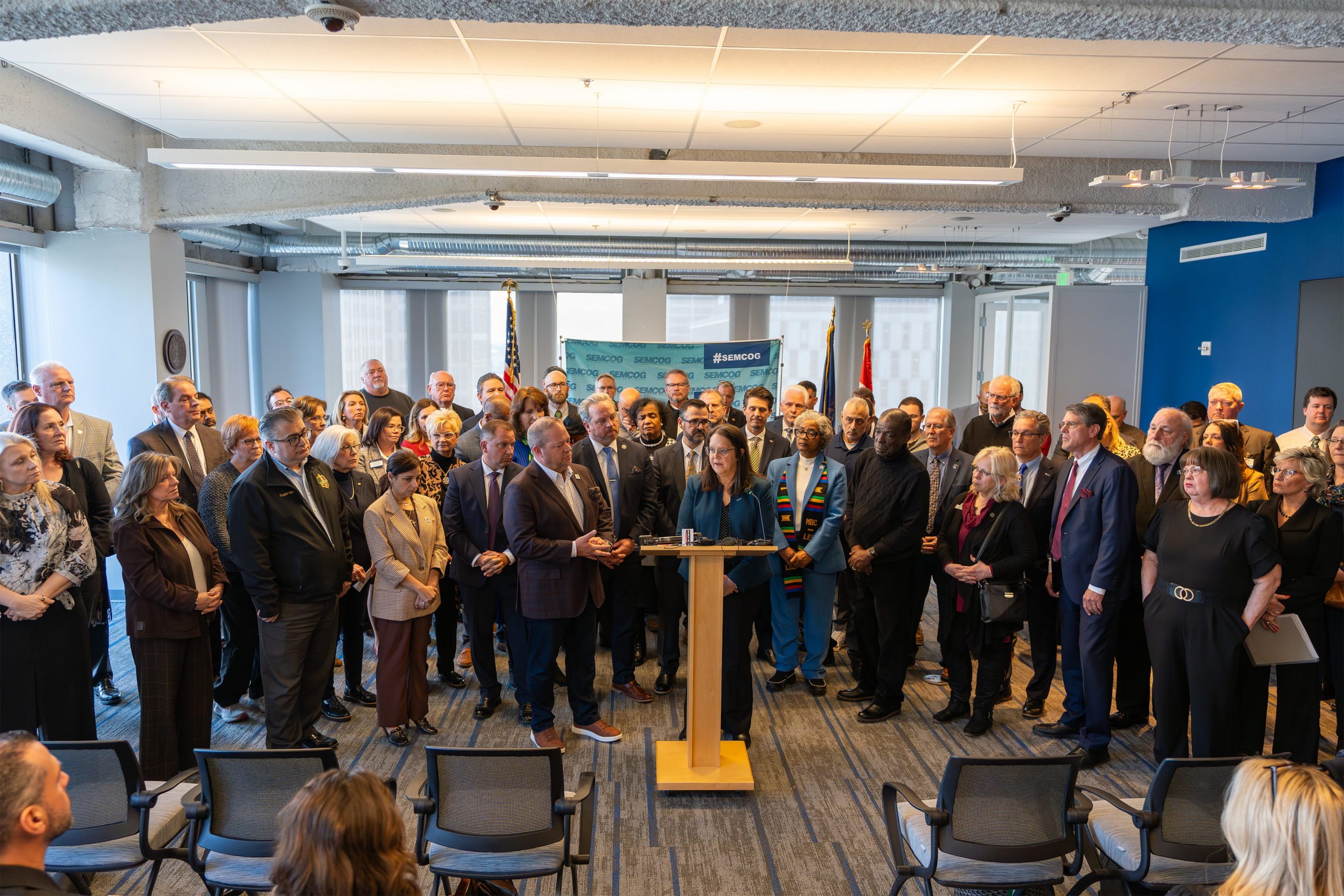A large group of people is gathered in a brightly lit room, standing and facing forward. A person is speaking at a podium, with several others standing closely behind. Flags and a banner featuring Salesforce logos are visible in the background. The room has rows of empty chairs in the foreground.