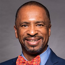 Close-up image of Scott Benson in a navy blue blazer, blue buton up and red polka dot bow tie. Against a grey background