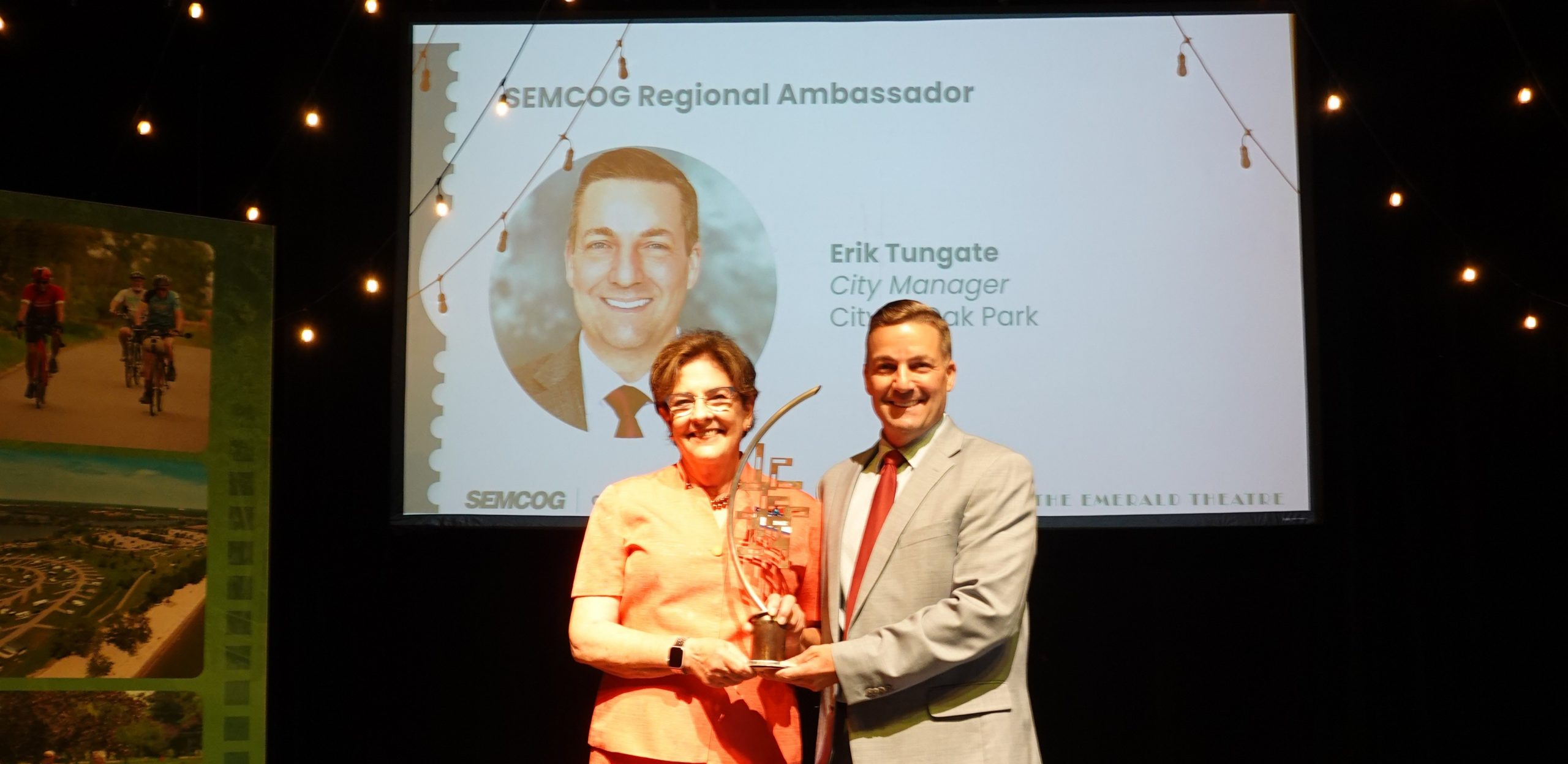 Two people present an award onstage at a SEMCOG event, standing before a presentation screen and audience.
