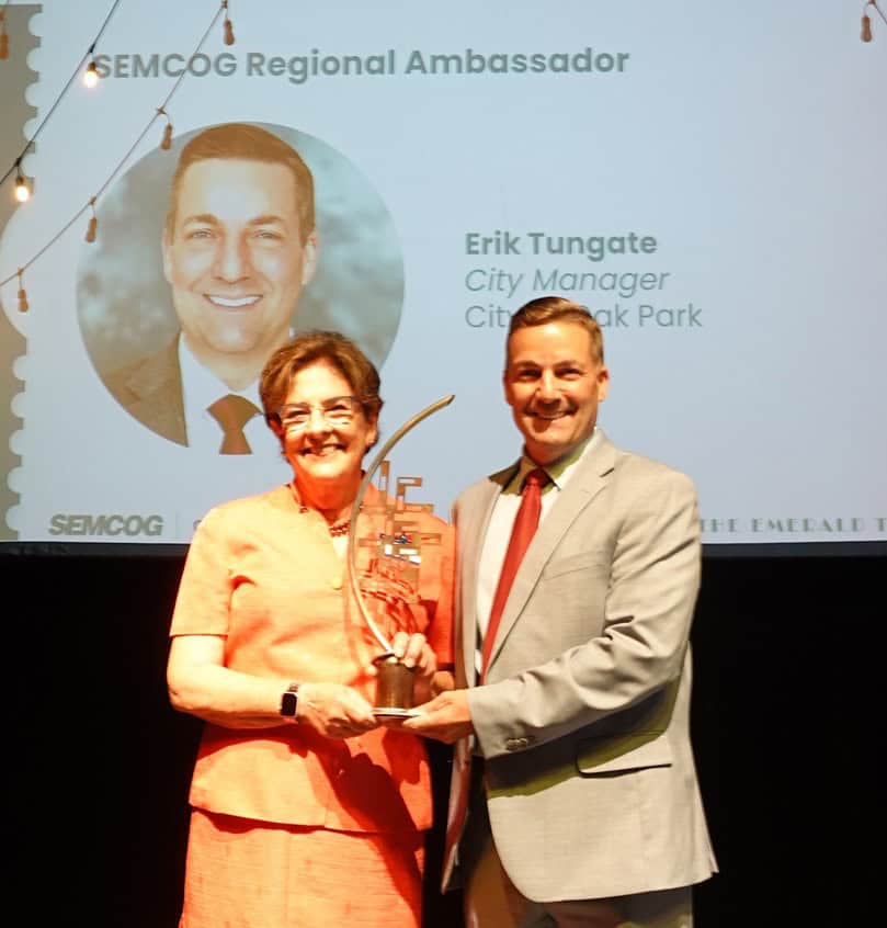 Two people present an award onstage at a SEMCOG event, standing before a presentation screen and audience.
