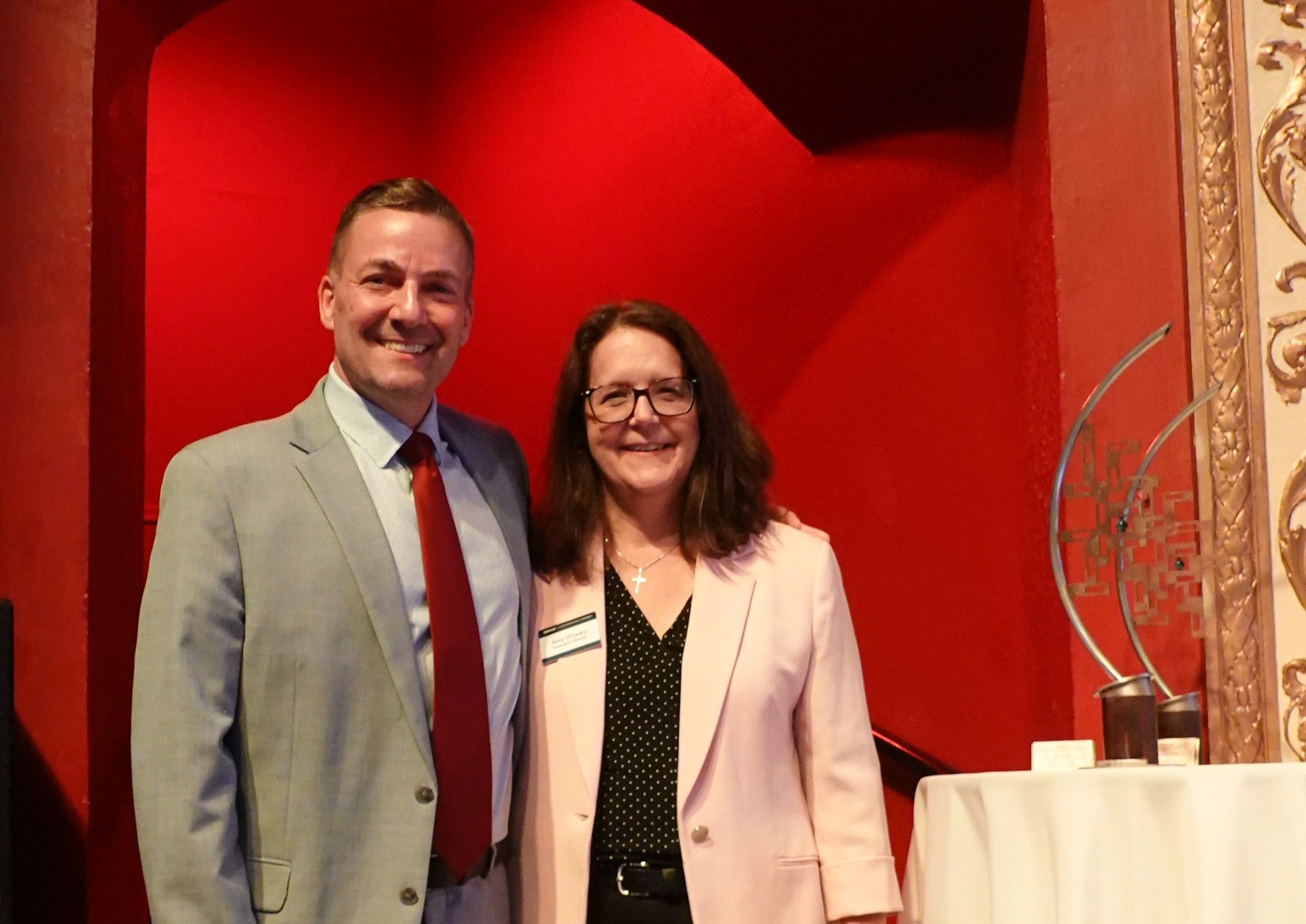 Two people in business attire standing together at an indoor event with red walls and a display table nearby.