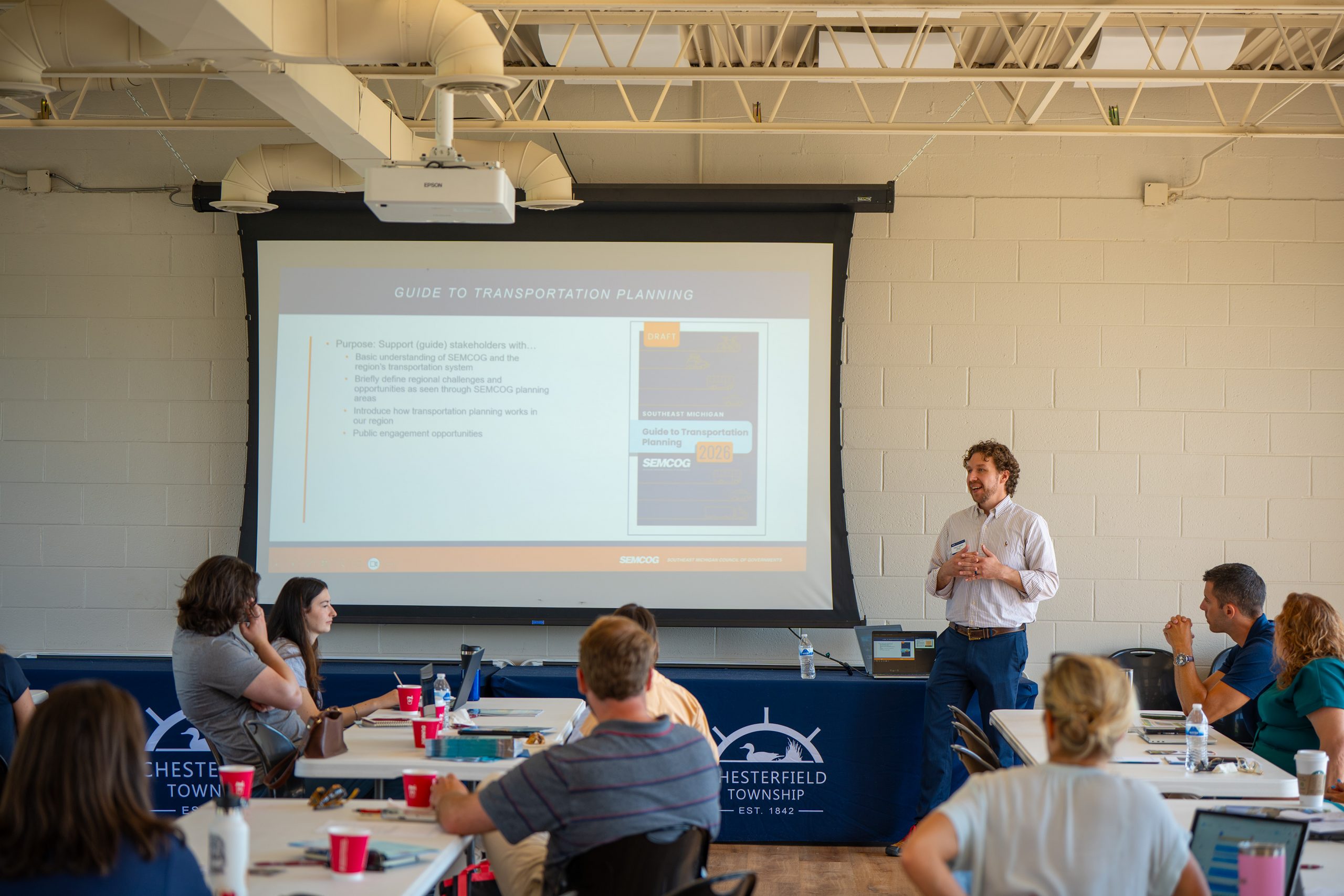 A speaker delivers a SEMCOG transportation planning presentation to workshop participants in a meeting room.