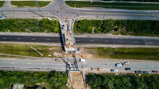 Aerial view of an active transportation infrastructure project where a new bridge or overpass is being constructed across a divided freeway. Construction equipment, work crews, temporary traffic control devices, and partially completed bridge spans are visible above multiple travel lanes. Adjacent roads, green space, and vehicles traveling through the corridor highlight a major roadway improvement project designed to enhance regional connectivity and transportation safety.