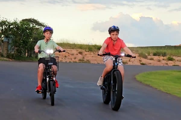 Two children wearing helmets ride bicycles on a paved path.