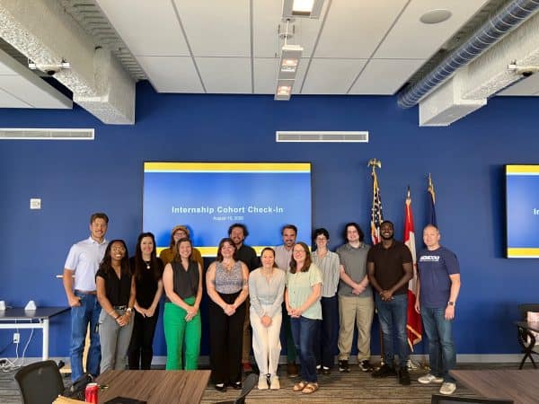 Group photo of interns and staff standing in a conference room in front of a presentation screen displaying 'Internship Cohort Check-In.'